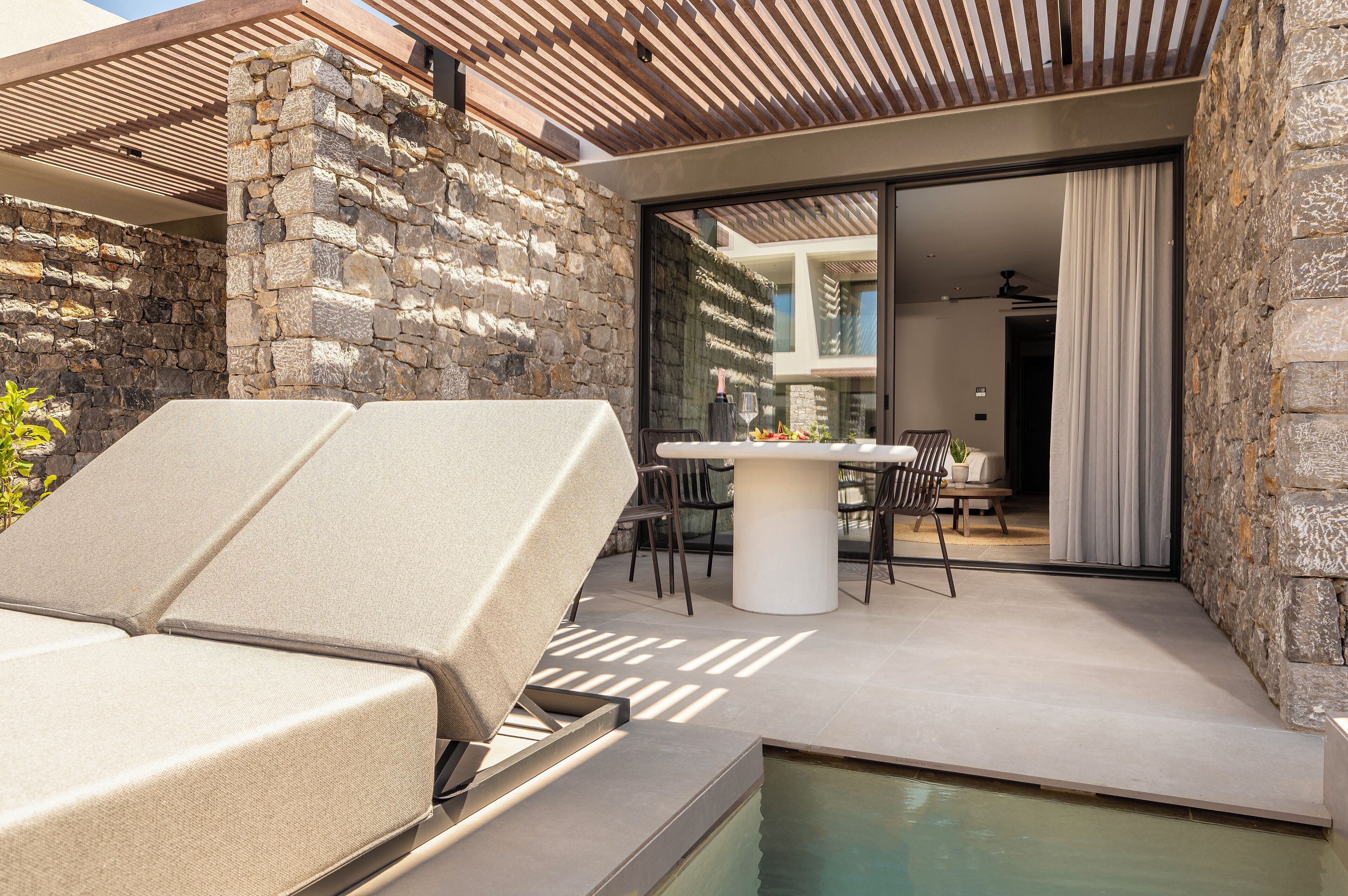 Zona junto a la piscina con una tumbona, paredes de piedra y un área de comedor con mesa y sillas.