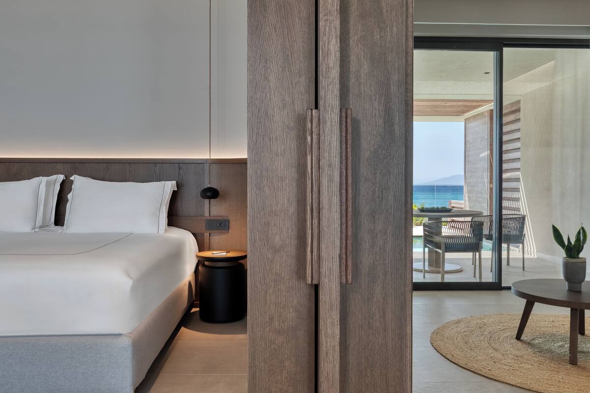 A modern bedroom with a large bed, a bedside table, and a view of a balcony with outdoor furniture.