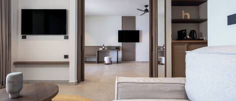 A modern hotel room with a large sofa, a small round table, a flat-screen TV mounted on the wall, a ceiling fan, and a kitchenette area.