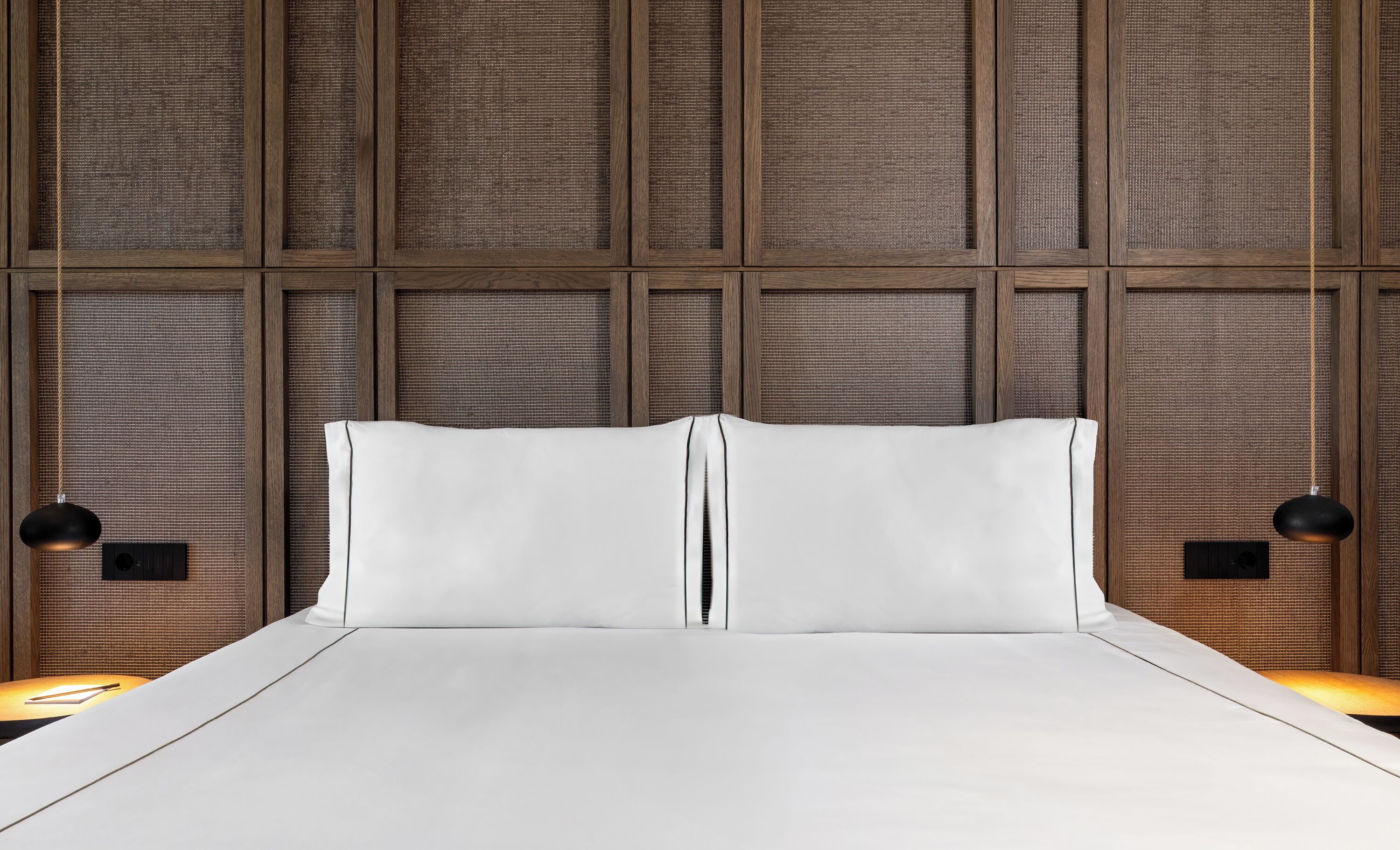 Una cama con sábanas blancas, cabecera de madera y dos lámparas negras fijadas a la pared.