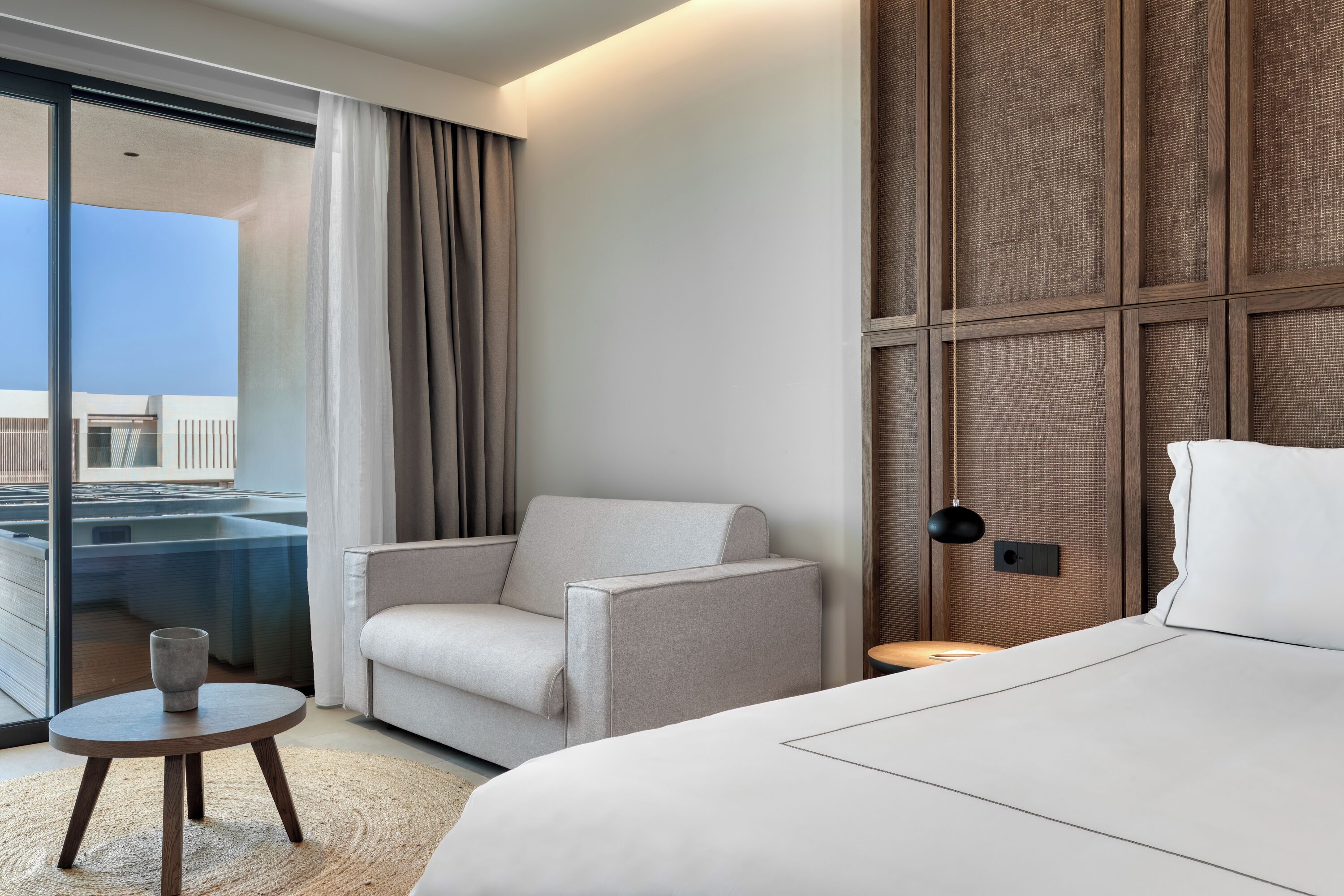 A hotel room with a bed, a chair, a small table, and a view of a pool and building through a glass door.