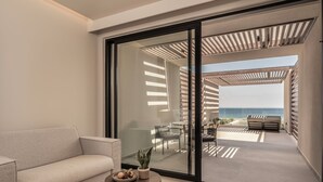 A modern living room with a sofa, a small round table with a plant and fruit bowl, and a sliding glass door leading to an outdoor patio with a view of the ocean. - Mosay All Suite Hotel - Adults friendly (Kos)