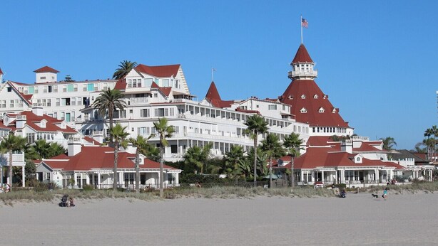 Ein großes, mehrstöckiges Hotel mit roten Dächern und weißen Wänden, gelegen an einem Strand mit Palmen.