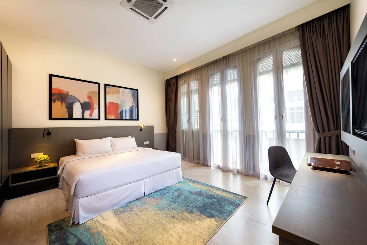 A modern hotel room with a large bed, a desk, and a TV. There are two framed pictures on the wall and a window with curtains.