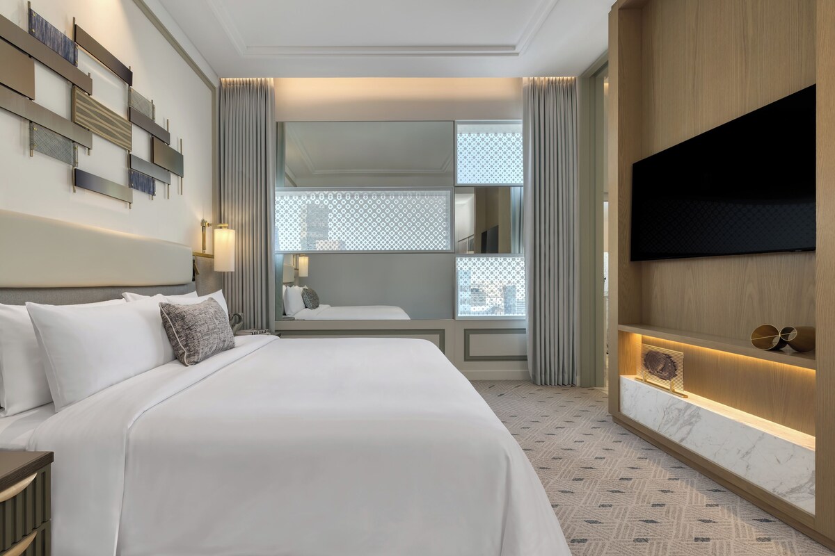 Habitación de hotel con una cama grande, un televisor de pantalla plana, un escritorio con lámpara y vistas a la ciudad a través de una ventana.