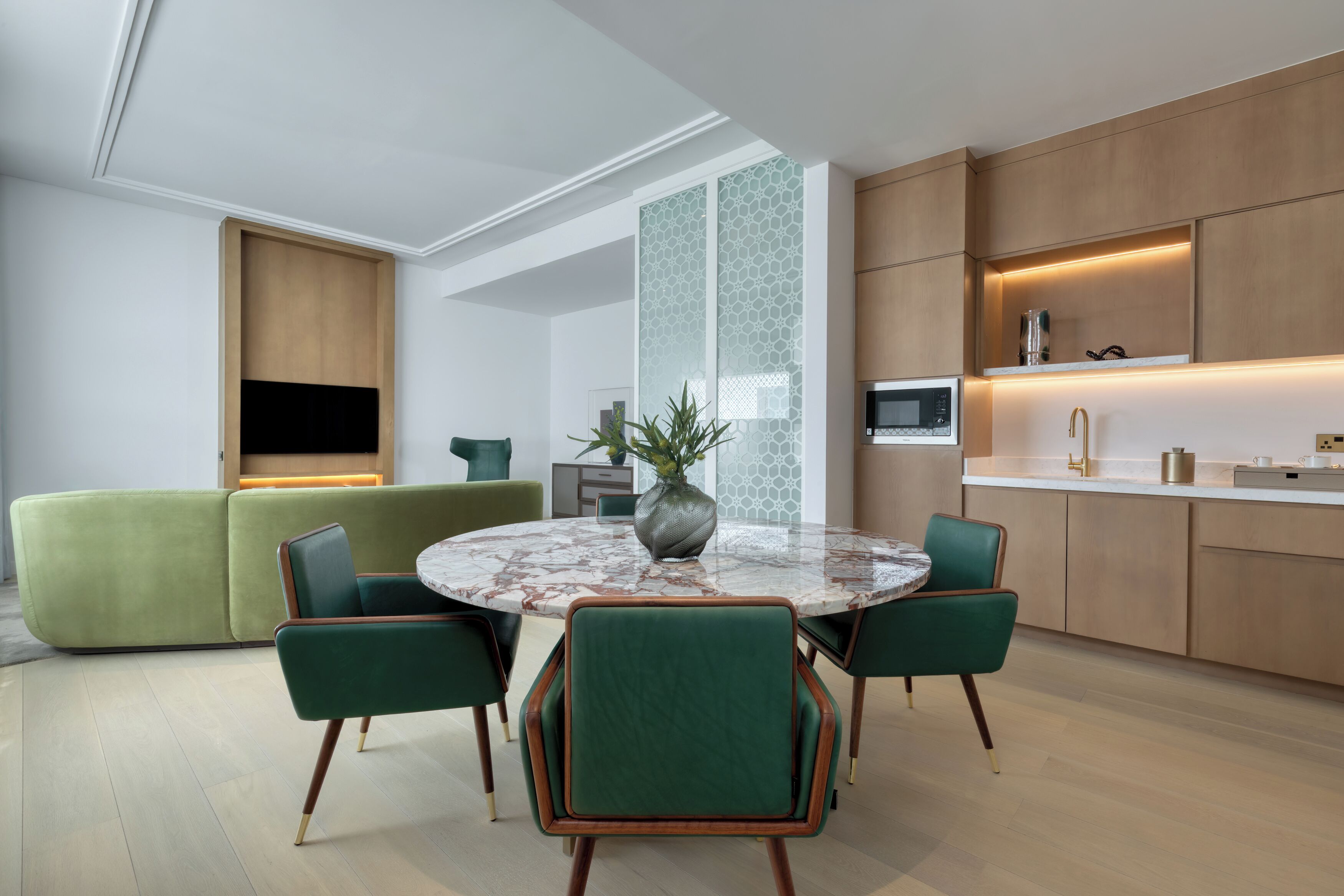 Un salon moderne avec une table ronde en marbre, des fauteuils à revêtement vert, un canapé vert et un coin cuisine doté de meubles en bois et d’un micro-ondes encastré.