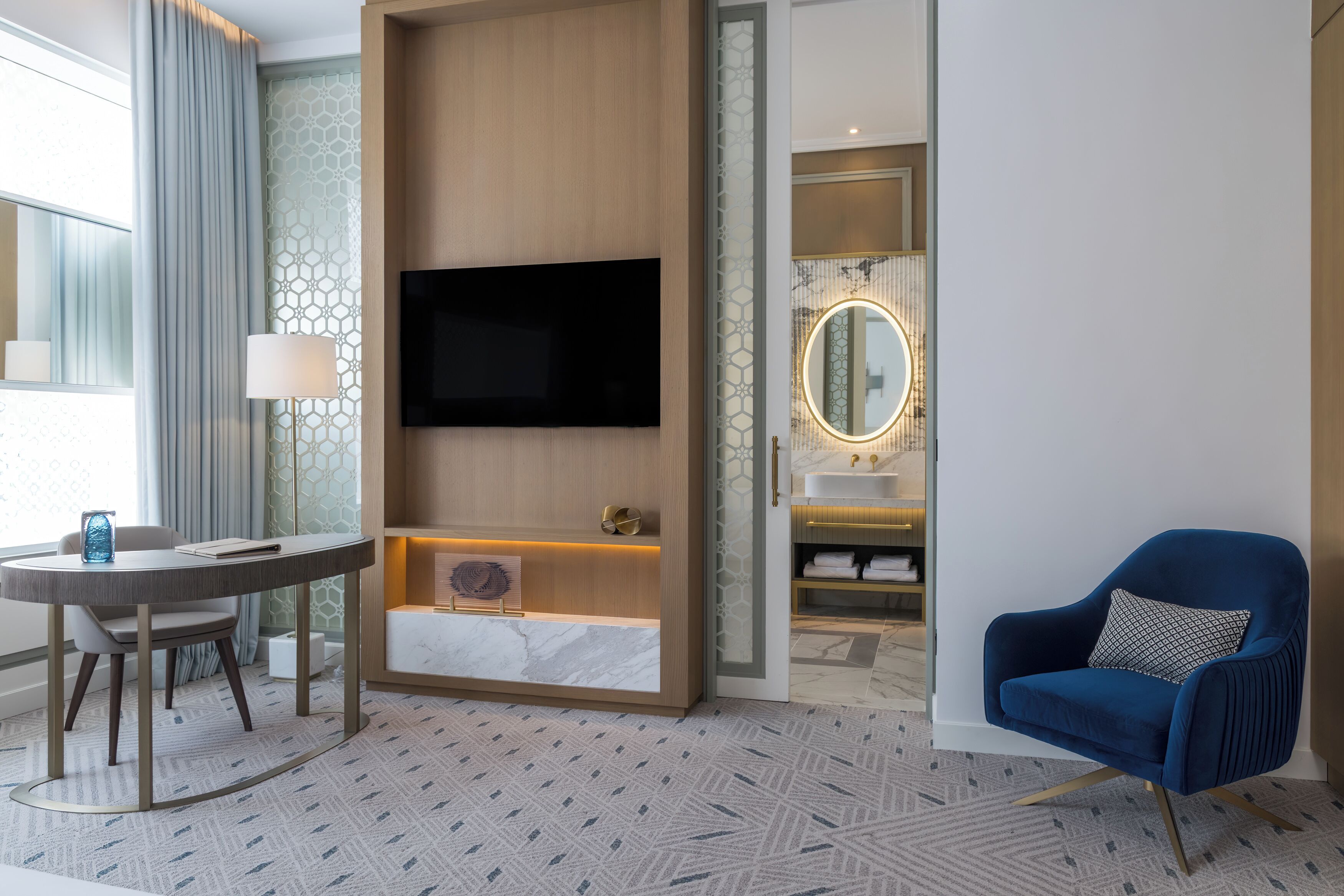 A modern hotel room with a flat-screen TV mounted on the wall, a round table with a lamp, a blue armchair, and a bathroom with a vanity and mirror.