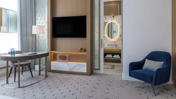 A modern hotel room with a flat-screen TV mounted on the wall, a round table with a lamp, a blue armchair, and a bathroom with a vanity and mirror.