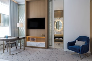 A modern hotel room with a flat-screen TV mounted on the wall, a round table with a lamp, a blue armchair, and a bathroom with a vanity and mirror. - Atheel KAFD Hotel (Riyadh)