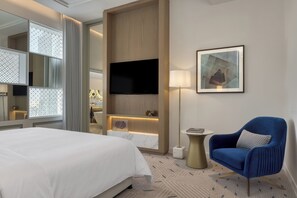 Habitación de hotel con una cama grande, un televisor de pantalla plana en la pared, un sillón azul, una pequeña mesa redonda y una obra de arte enmarcada en la pared.
