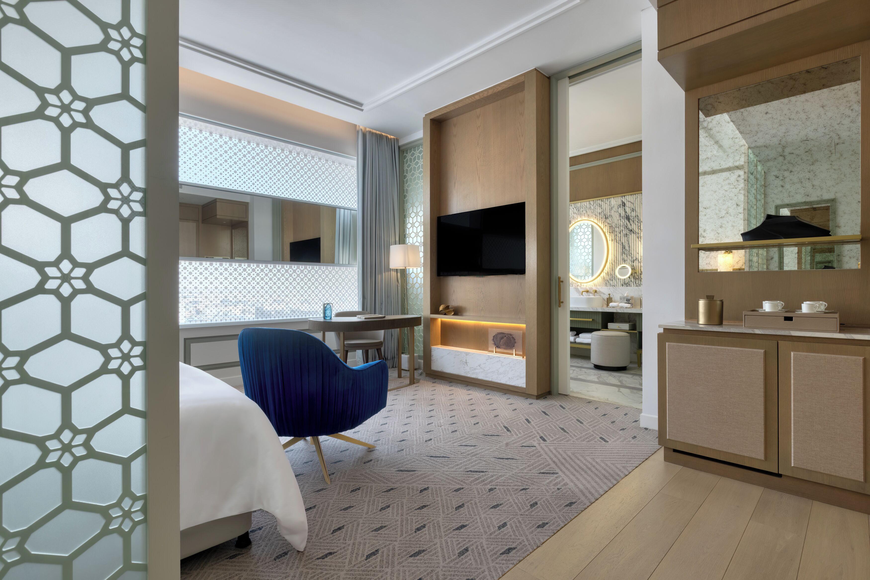 Habitación de hotel con cama, escritorio, silla, televisor y baño con espejo, además de una alfombra estampada.
