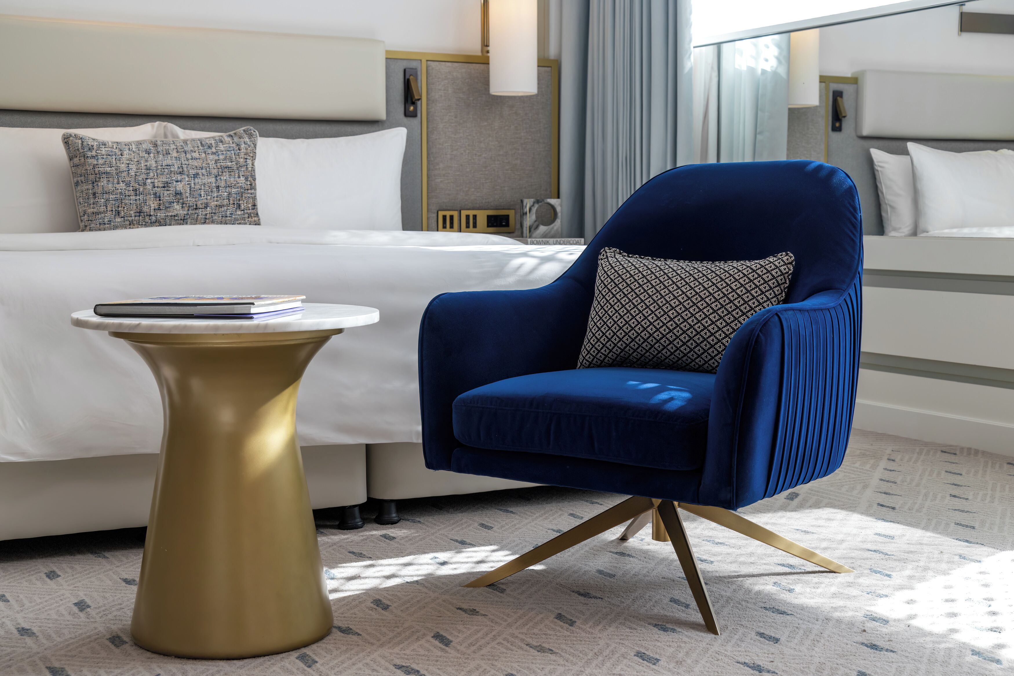 A hotel room with a bed, a blue armchair, a small round table, and a patterned pillow.
