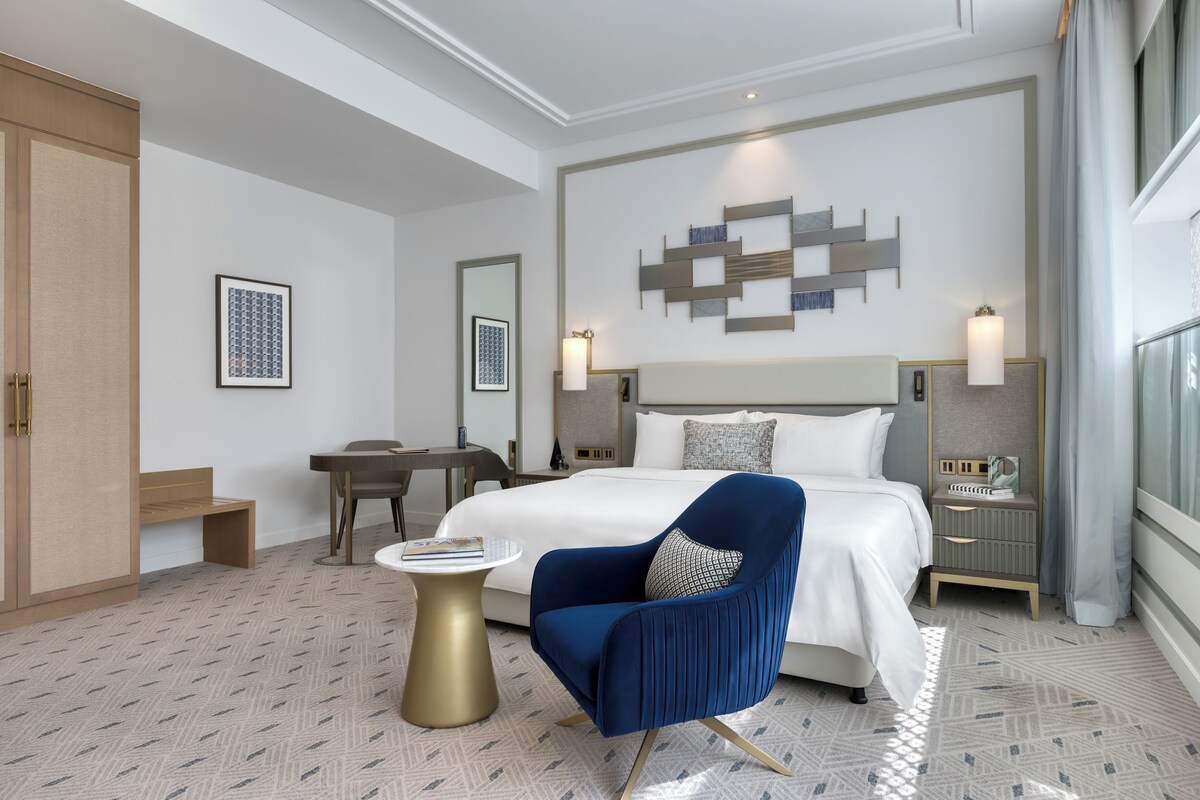Una habitación de hotel con cama, escritorio, silla, mesita de noche y lámpara de pared.