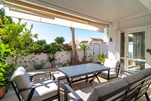 Une terrasse extérieure avec une table et des chaises, entourée de plantes en pot et d’un arbre.