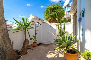 Une cour étroite agrémentée de plantes en pot, d’un arbre et d’un bâtiment blanc surmonté d’un toit en tuiles.