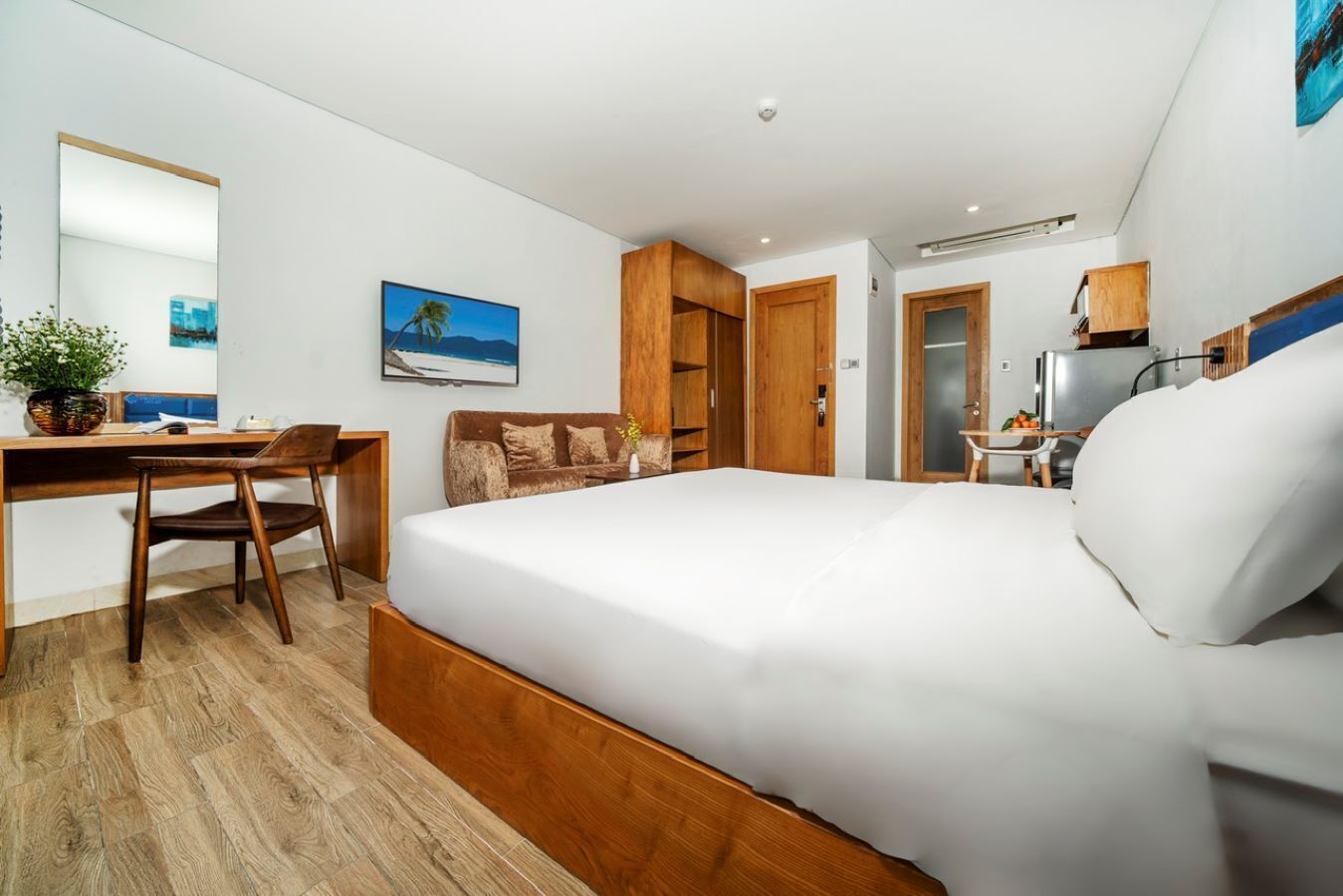 A modern hotel room with a large bed, a desk, a chair, a small table, and a wardrobe.