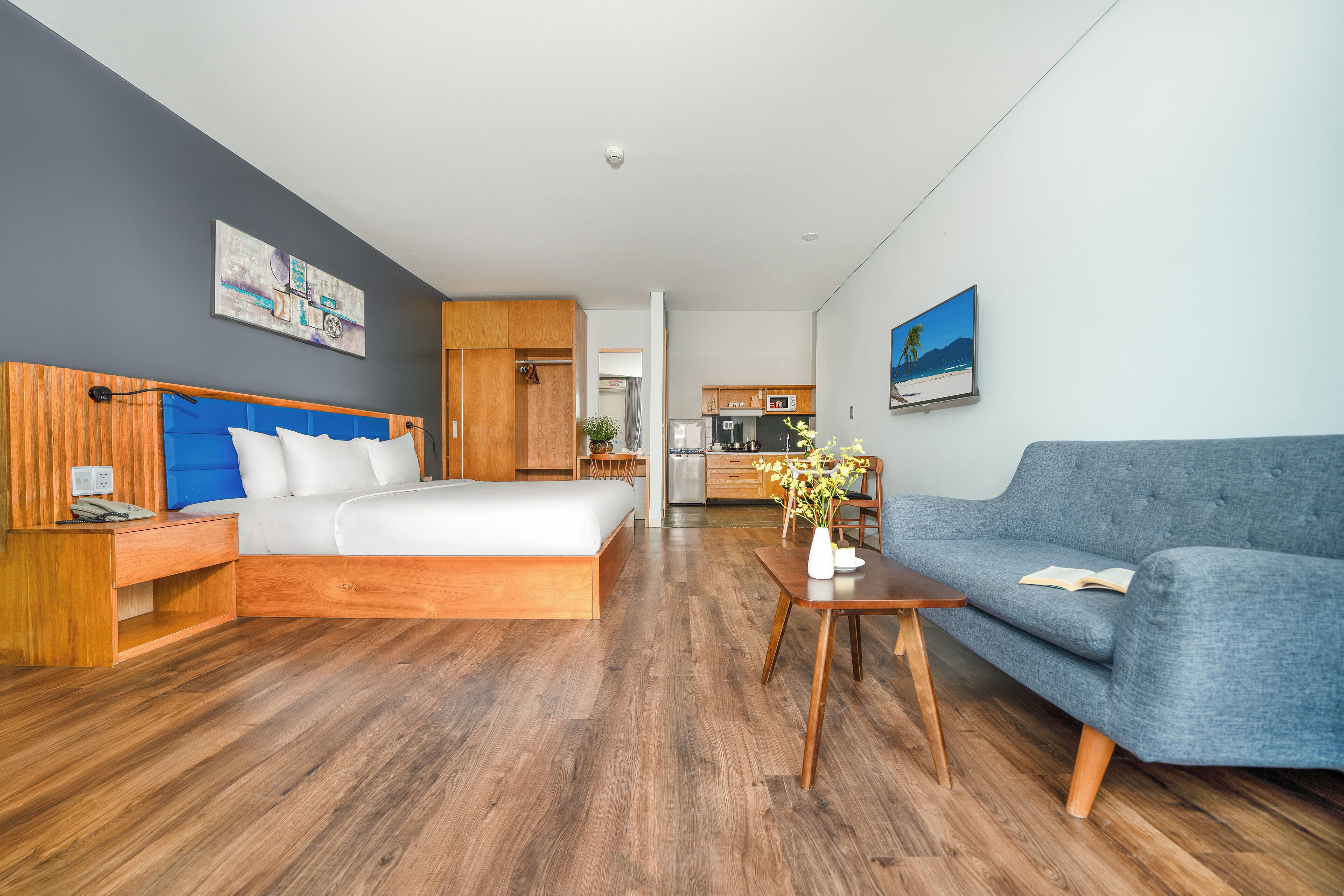 Una habitación de hotel moderna con piso de madera, una cama con cabecera azul, un sofá gris, una mesita con un florero y una cocina al fondo.