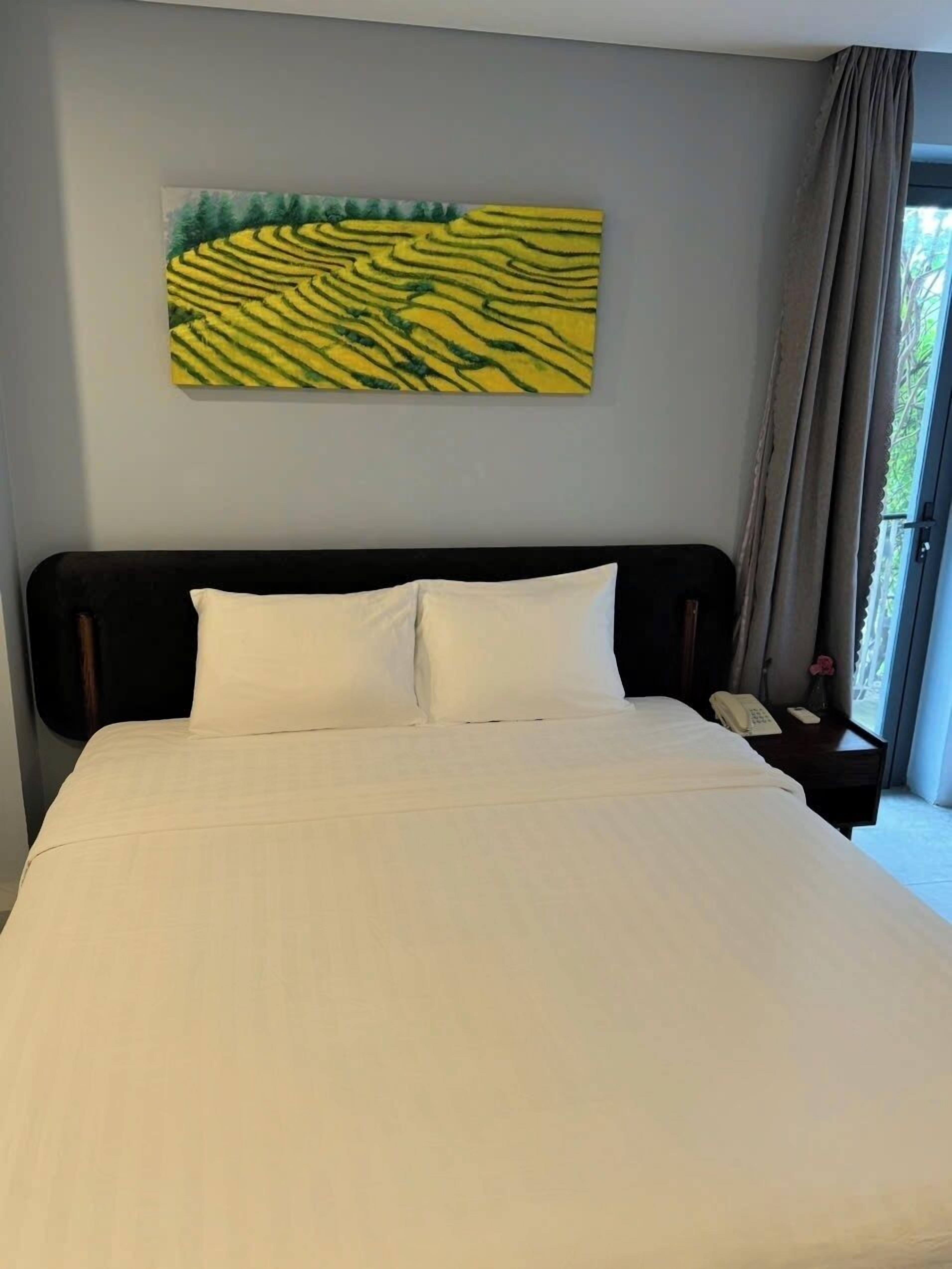 Habitación de hotel con una cama grande, una mesita de noche con teléfono y un florero, y una ventana con cortinas.