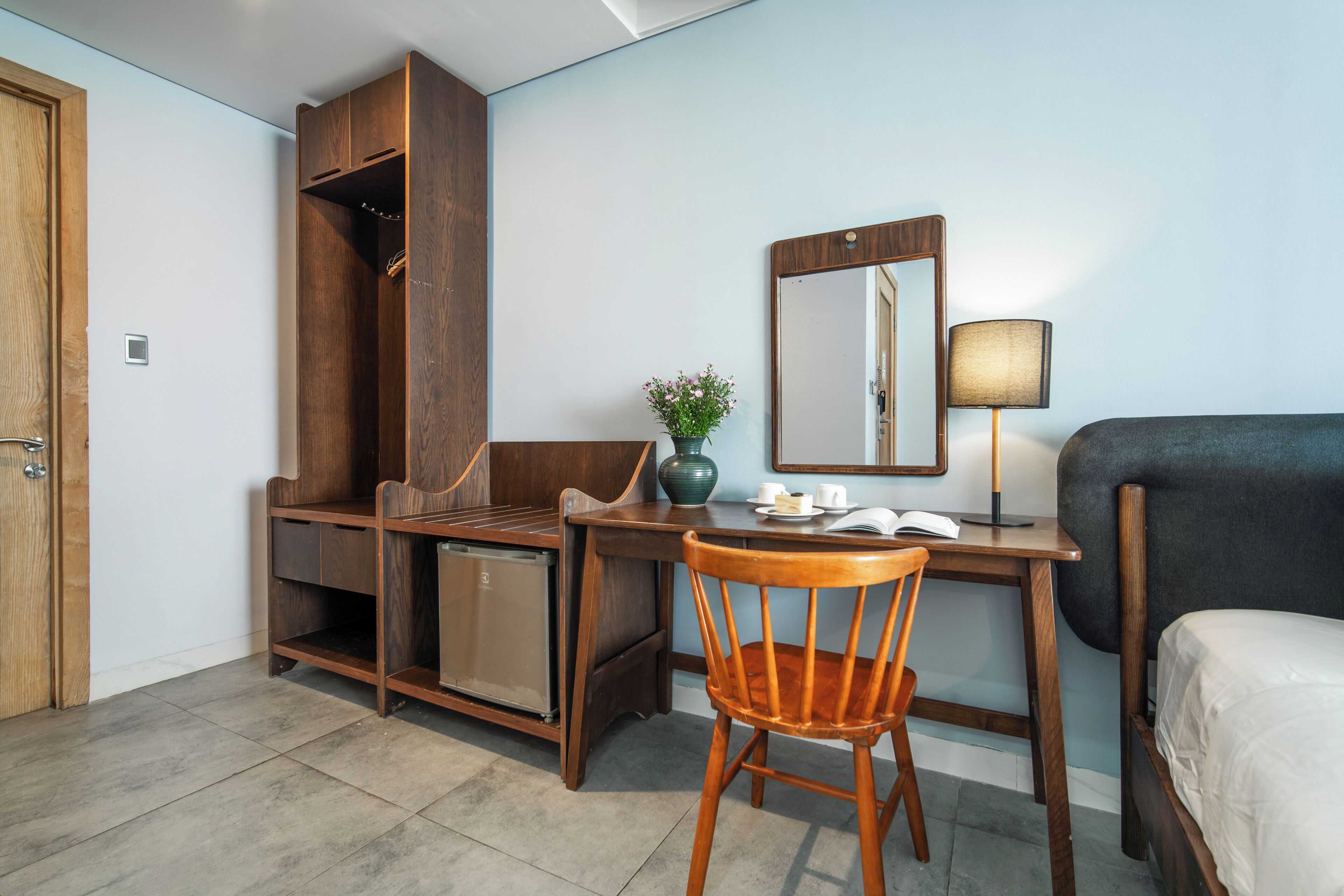 Una habitación de hotel compacta con un escritorio de madera, una silla, una cama, un armario, un refrigerador pequeño, un espejo y una lámpara de noche.