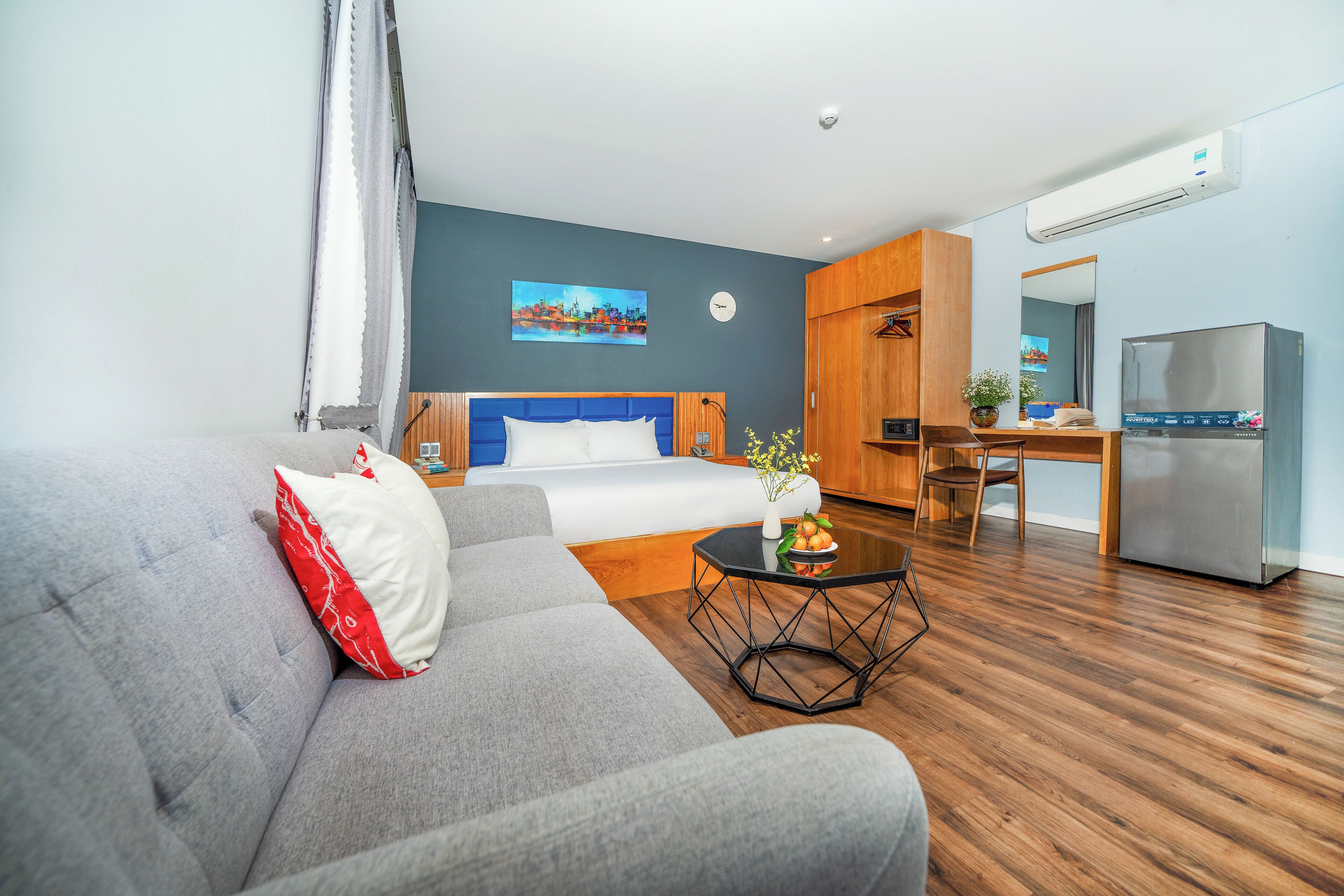 A modern hotel room with a grey sofa, a wooden dining table, a small kitchen area, and a painting on the wall.
