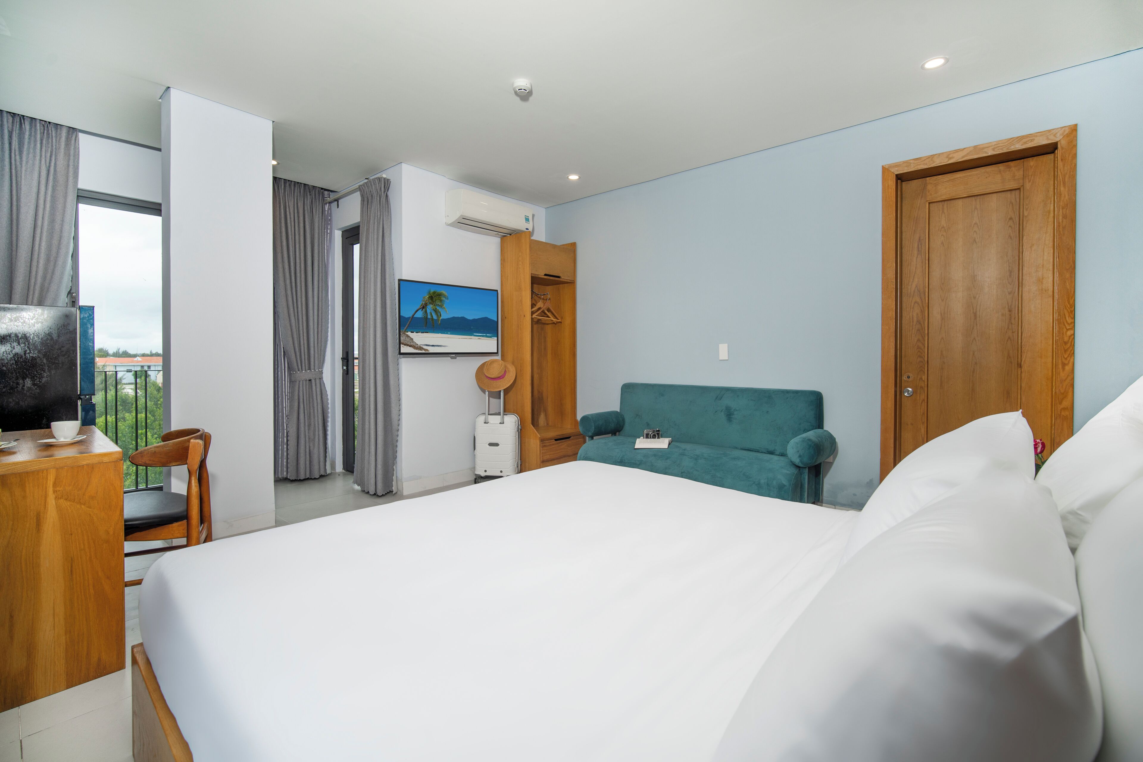 Quarto de hotel com uma cama grande, escrivaninha com televisão, janela com cortinas, ar condicionado fixado na parede e um quadro de uma praia.