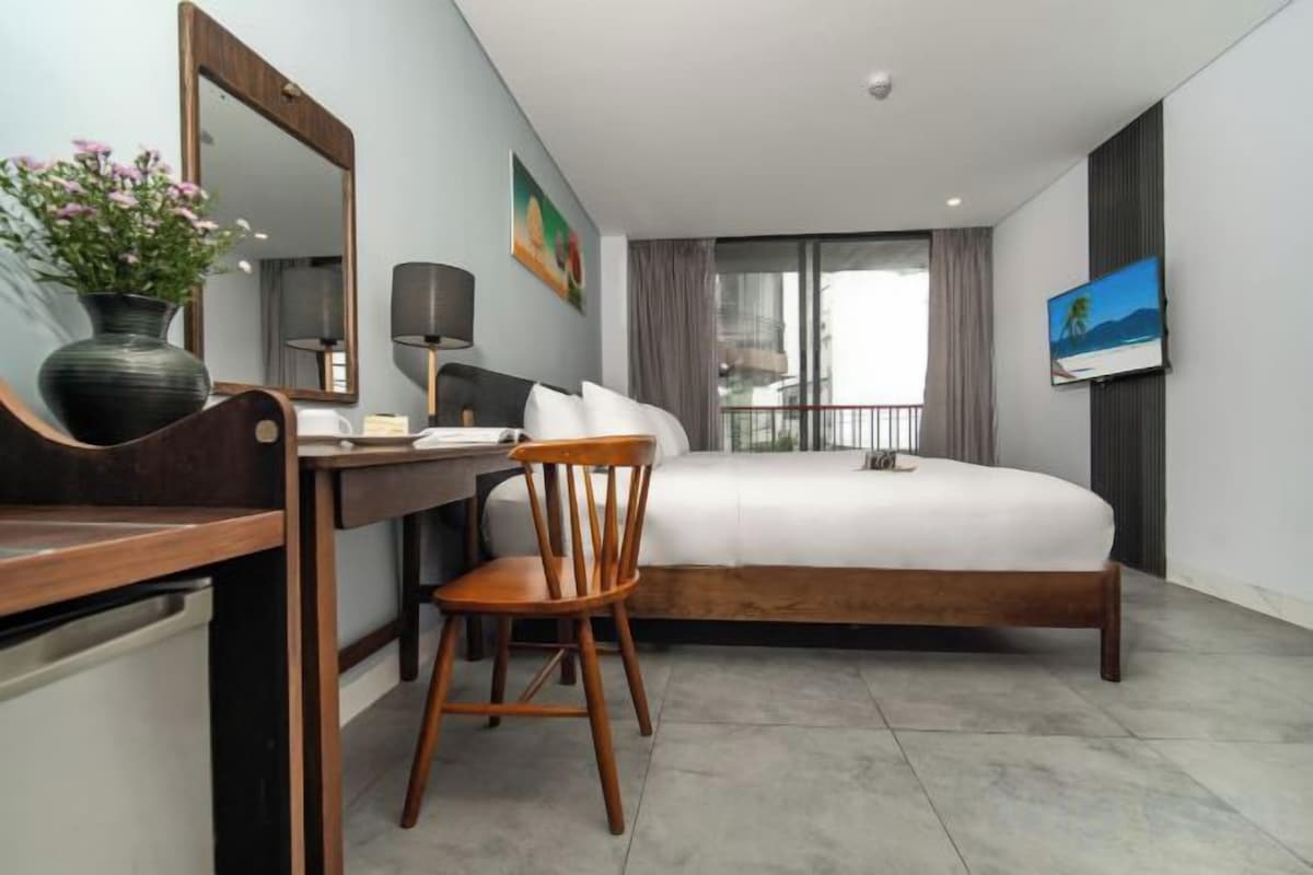 Habitación de hotel con cama, escritorio con silla, espejo, televisor y ventana con cortinas.