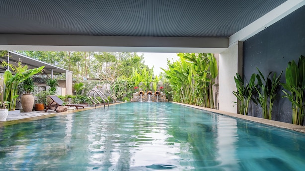 A swimming pool with a stone wall and potted plants on one side, and a covered area with lounge chairs on the other.
