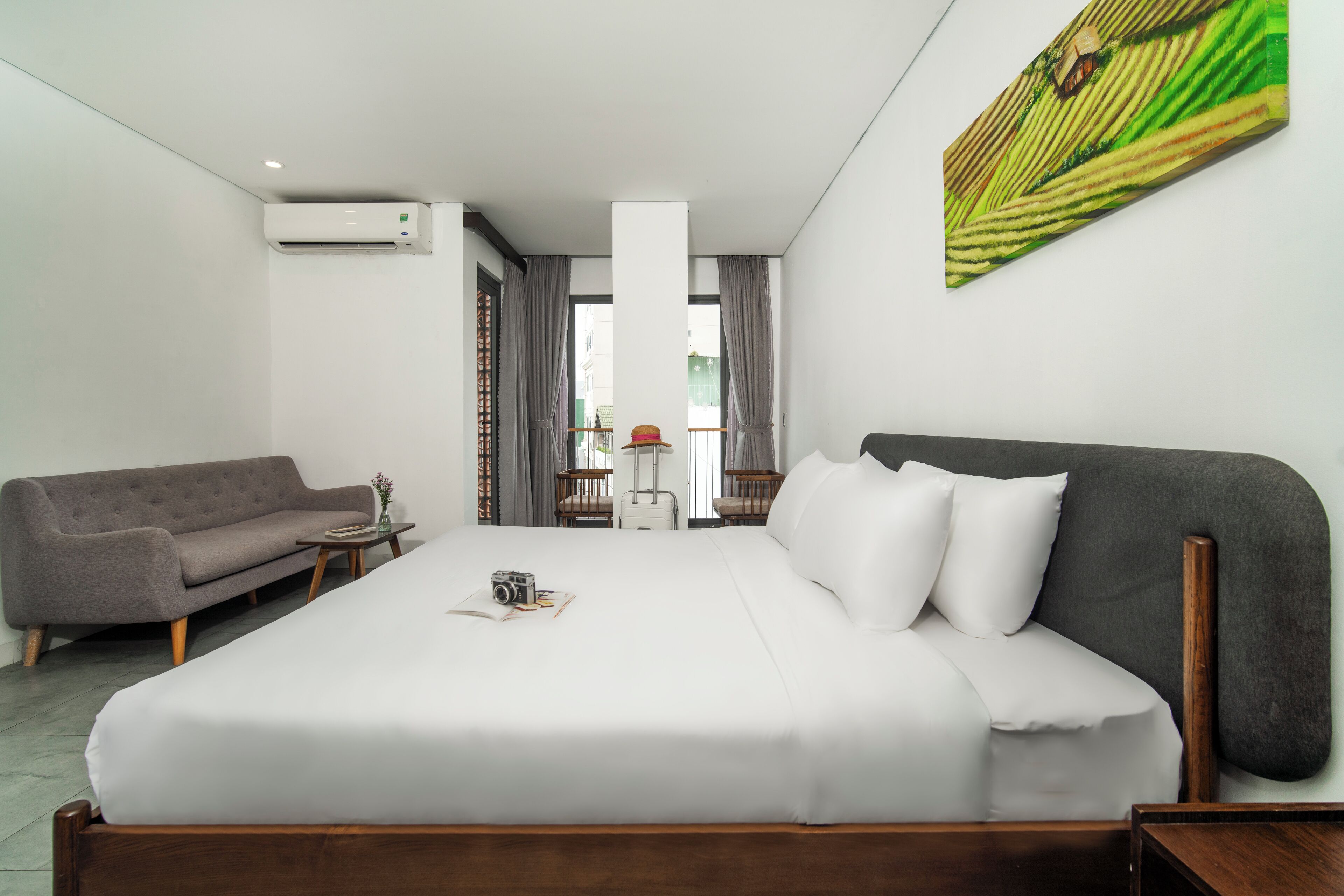 A hotel room with a large bed, a sofa, a small table, and an air conditioning unit.
