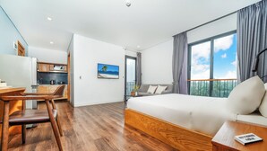 A modern hotel room with a wooden floor, a large bed, a sofa, a desk, and a kitchen area.