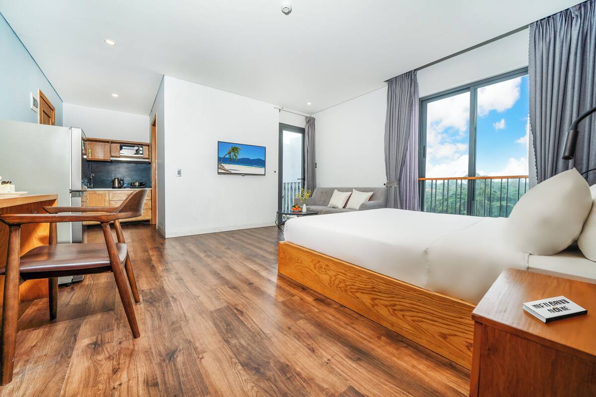 Moderni hotellihuone, jossa on puulattia, suuri sänky, sohva, työpöytä ja pieni keittiö.