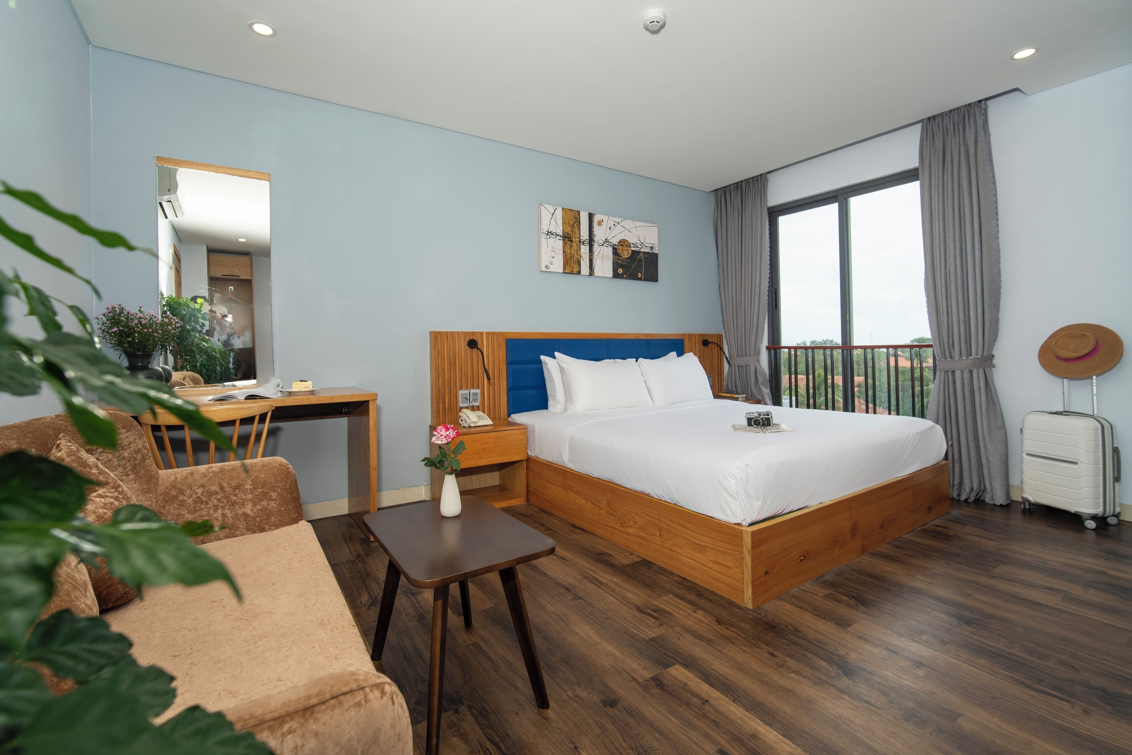 A modern hotel room with a large bed, a desk, a chair, a small table, and a balcony with curtains.