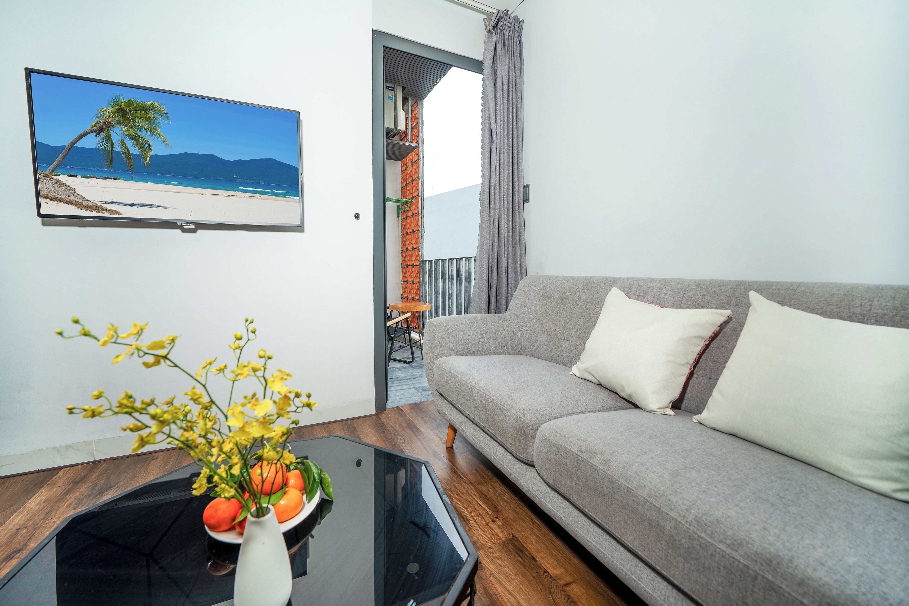 A living room with a grey sofa, a black coffee table with a white vase and yellow flowers, and a TV displaying a beach scene.