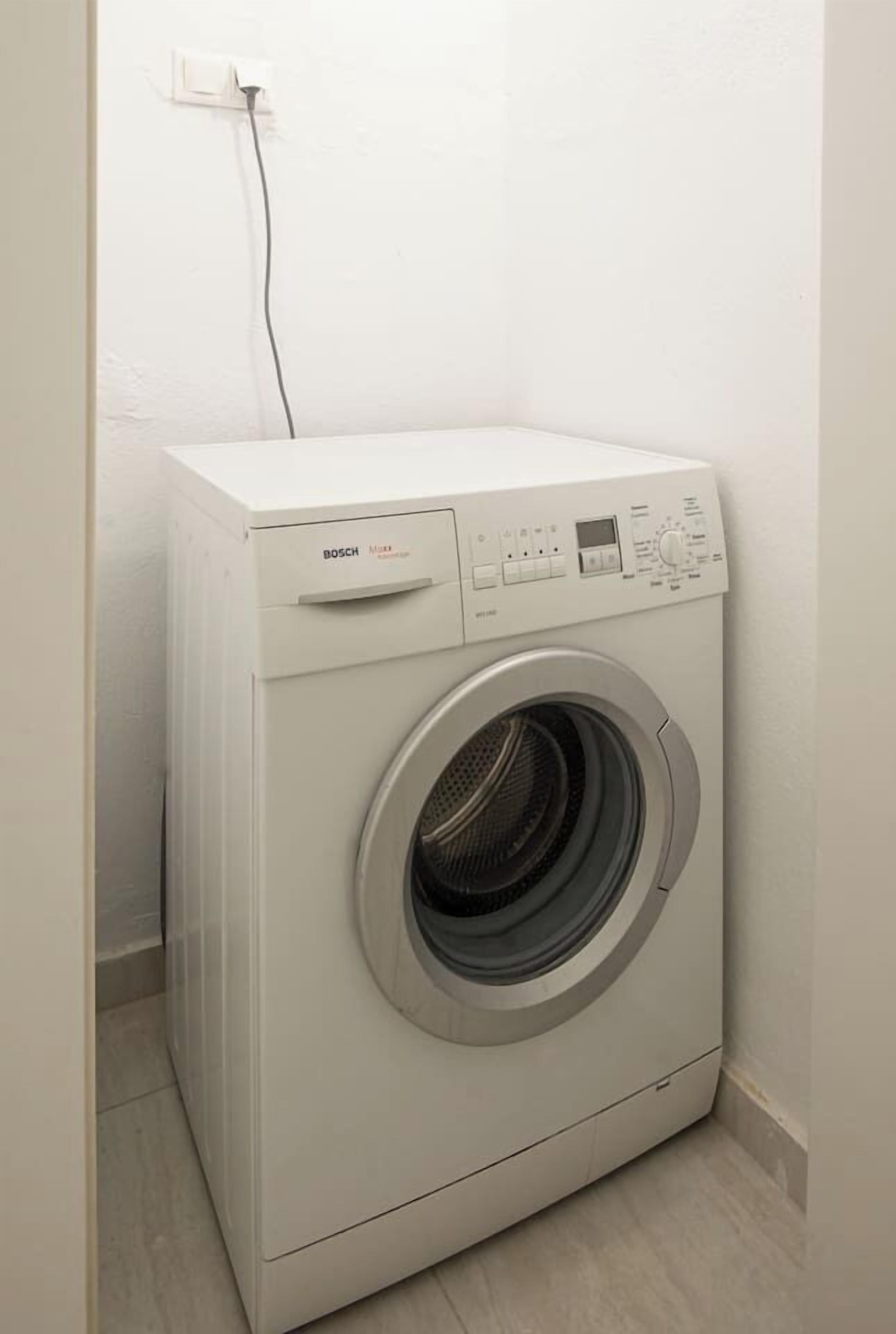 Een Bosch wasmachine in een hoek van een kamer met een witte muur en een zichtbare elektriciteitsstekker.