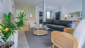 A modern living room with a grey sofa, white armchairs, and a round coffee table. There is a kitchen area in the background with a large window. - w*| Sublime 2BR w/ Rooftop In Casco Antiguo (Panamá)