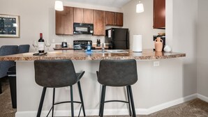 A modern kitchen with granite countertops, dark wooden cabinets, a black refrigerator, and a microwave. There are two bar stools and a wine bottle on the counter. - New Albany 2BR | 2-Car Garage | Walk to Dinner (Columbus)
