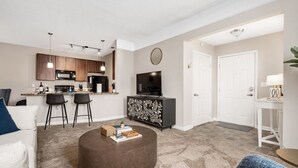 A modern living room with a round coffee table, a sofa, and a TV. There is a kitchen area with a microwave and a dining table with chairs. - New Albany 2BR | 2-Car Garage | Walk to Dinner (Columbus)