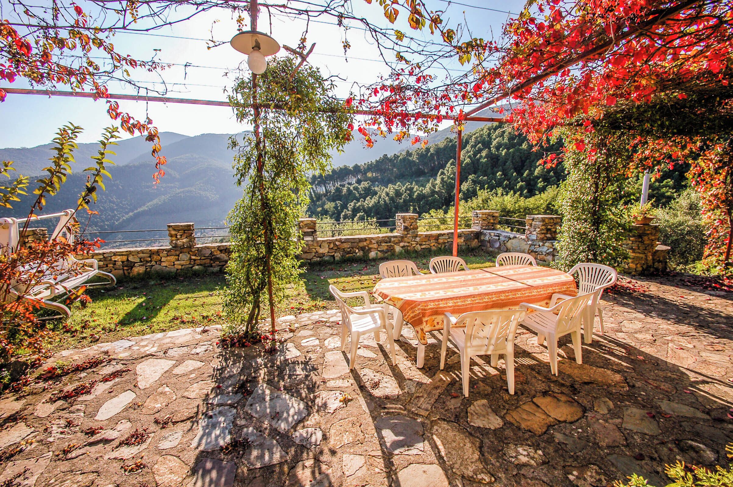 A stone patio with a table and chairs, surrounded by greenery and a stone wall.