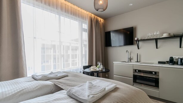 A hotel room with a bed, a small table with a chair, a kitchenette, a TV mounted on the wall, and a large window with sheer curtains. - Frost Apartments - Economy Studio (Reykjavík)