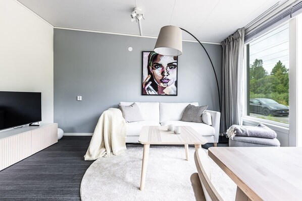 Un salon moderne avec un canapé blanc, un téléviseur à écran plat, une table basse et une grande fenêtre donnant sur un paysage verdoyant et une voiture.