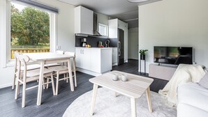 A modern kitchen with white cabinets, a dining area with a wooden table and chairs, a living room with a sofa and a TV, and a large window with a view of trees. - Private Chalet (Skelleftea)