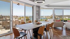 Un vaste espace de salle à manger avec une grande table et des chaises, de grandes fenêtres offrant une vue sur la ville et la mer, et un luminaire moderne au plafond.