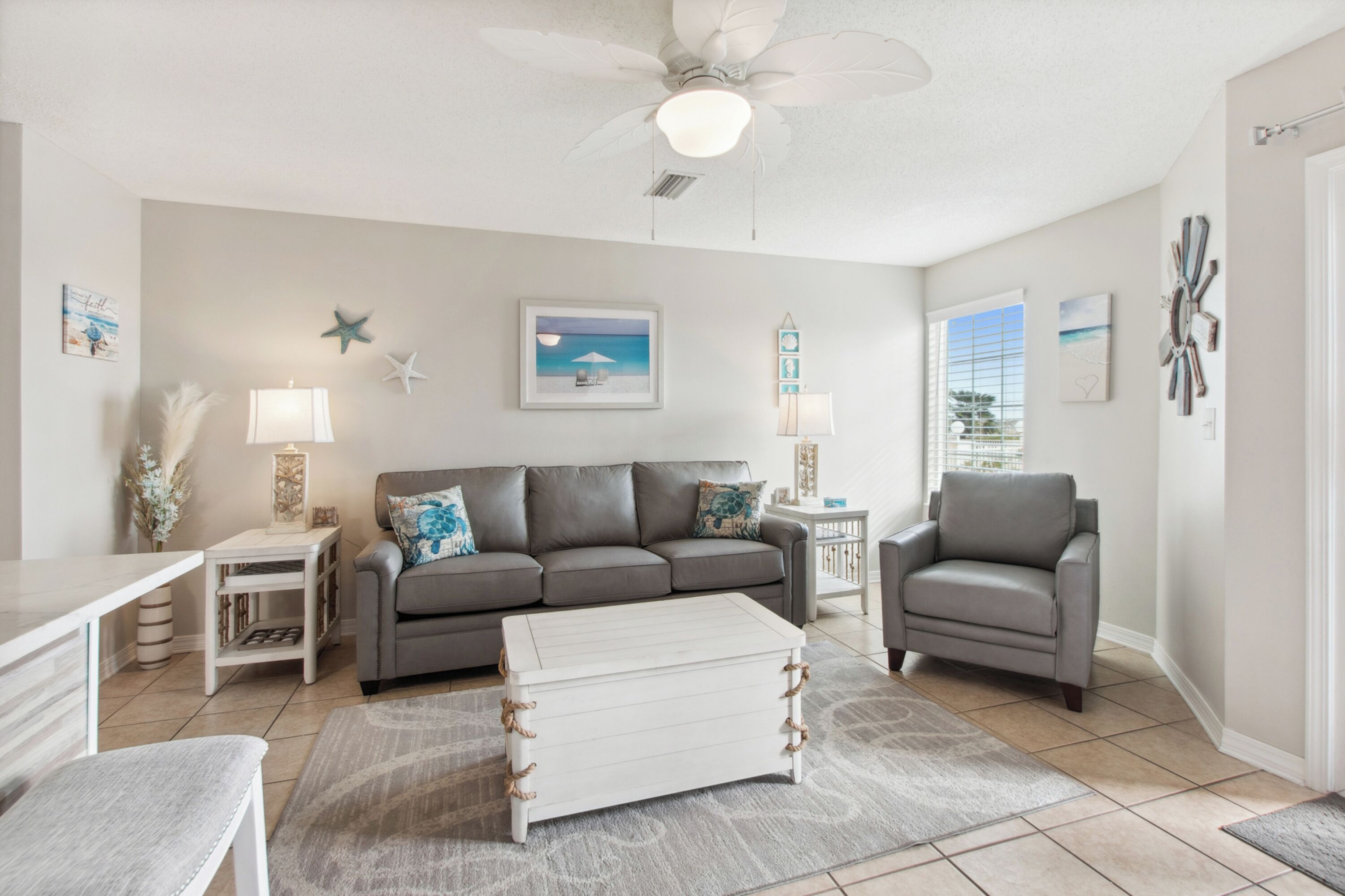 A living room with a gray sofa, a white coffee table, a gray armchair, and a ceiling fan. There are wall decorations including a starfish, a framed picture, and a clock.