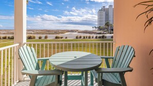 Een balkon met twee groene Adirondack-stoelen en een tafeltje, met uitzicht op een strand en een gebouw in de verte.