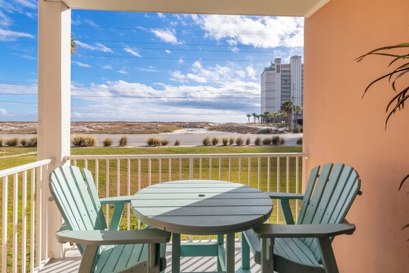 Un balcón con dos sillas Adirondack verdes y una pequeña mesa, con vista a una playa y un edificio lejano.