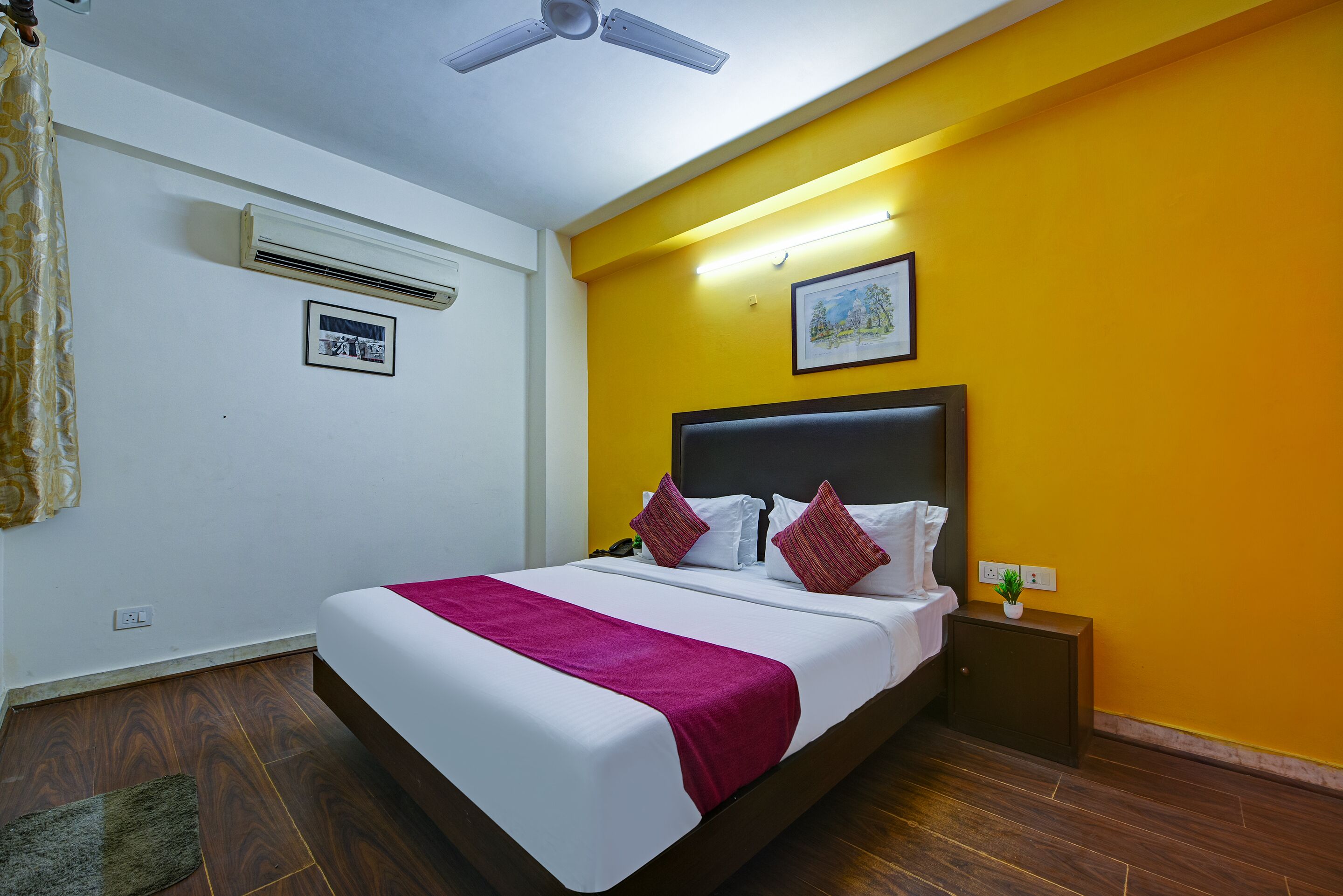 Habitación de hotel con cama, mesita de noche, aire acondicionado y ventilador de techo, además de un cuadro en la pared.