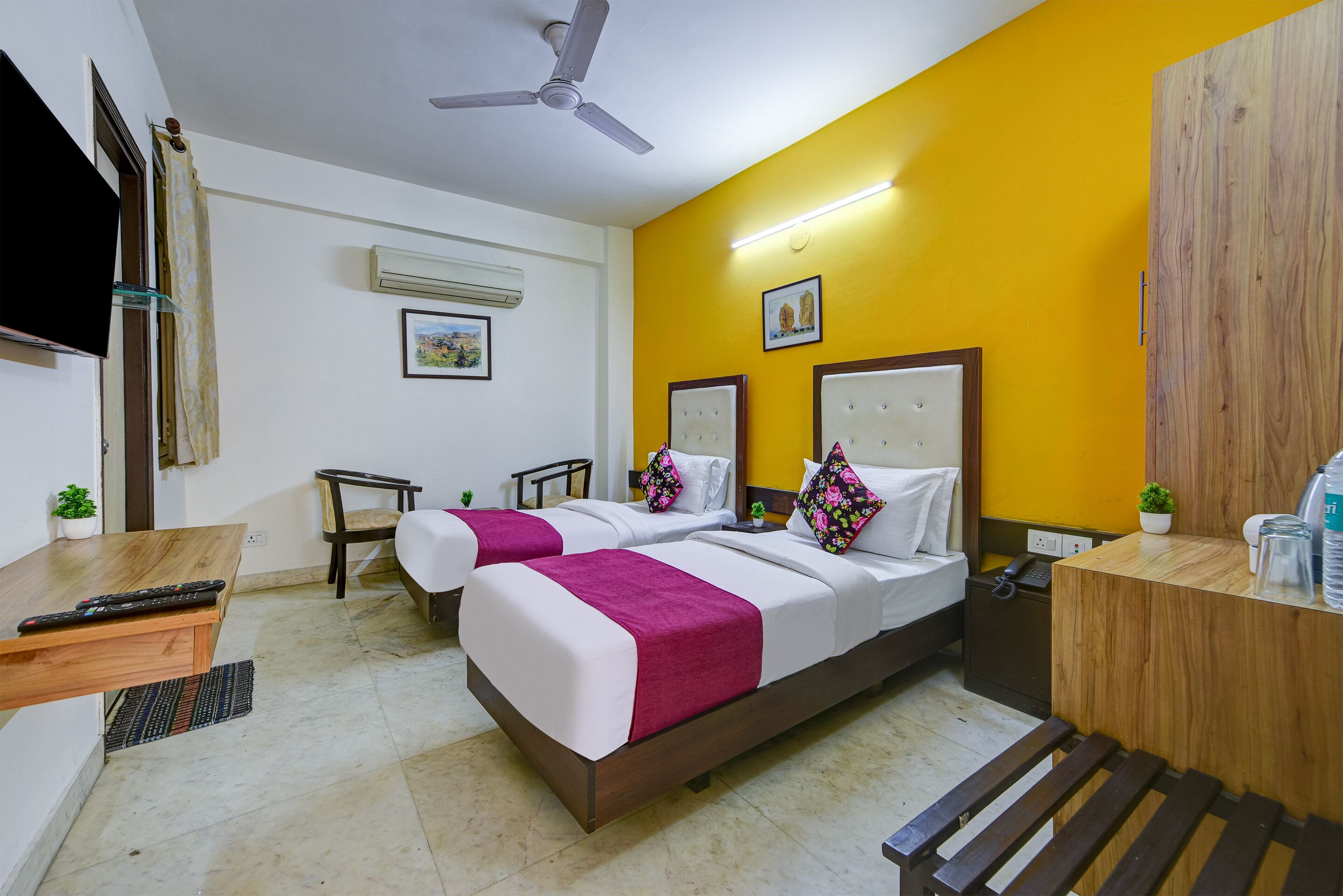 A hotel room with two beds, a desk, a chair, a ceiling fan, and a TV.