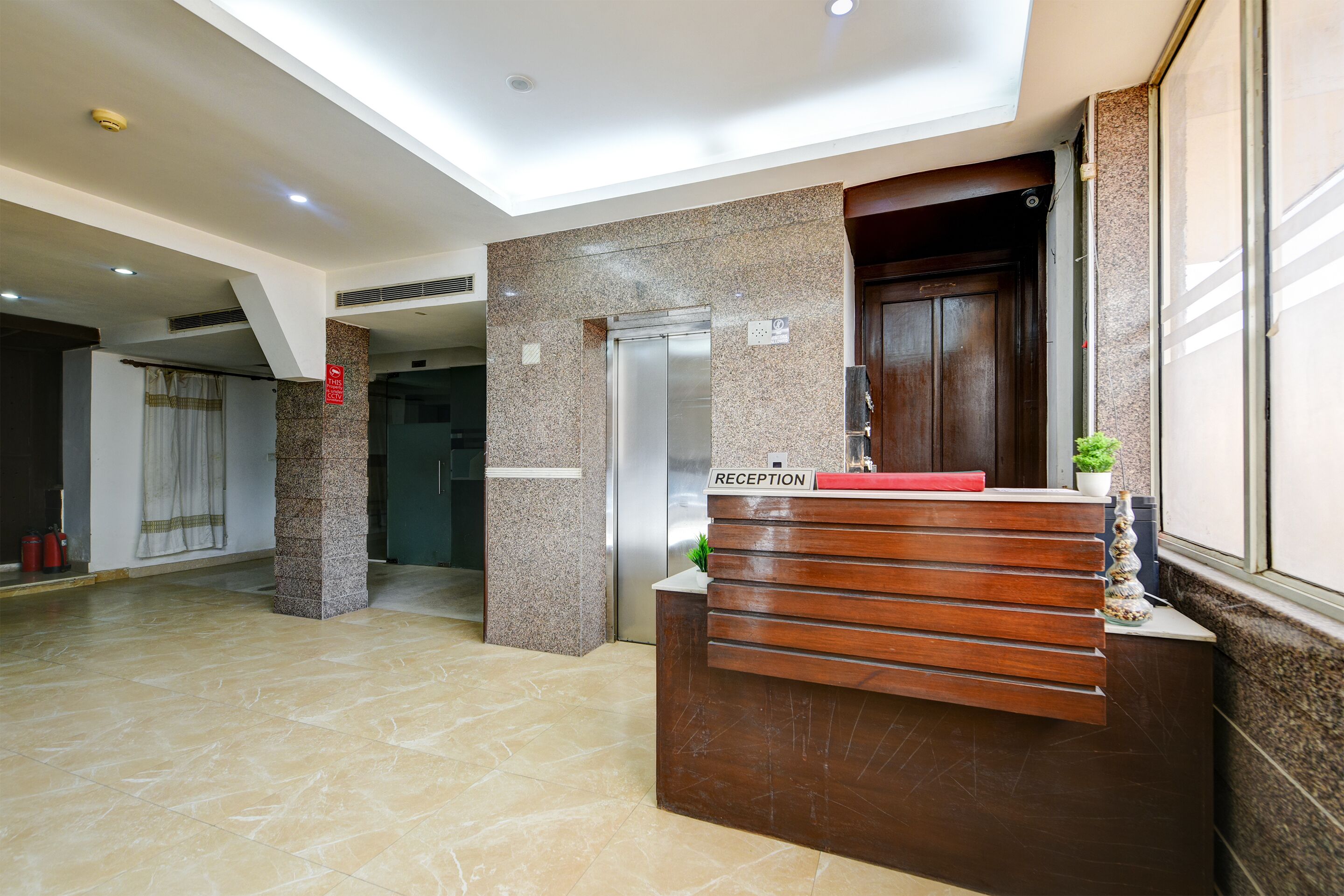 A hotel reception area with a reception desk, a reception sign, a wall-mounted elevator, and a fire extinguisher.
