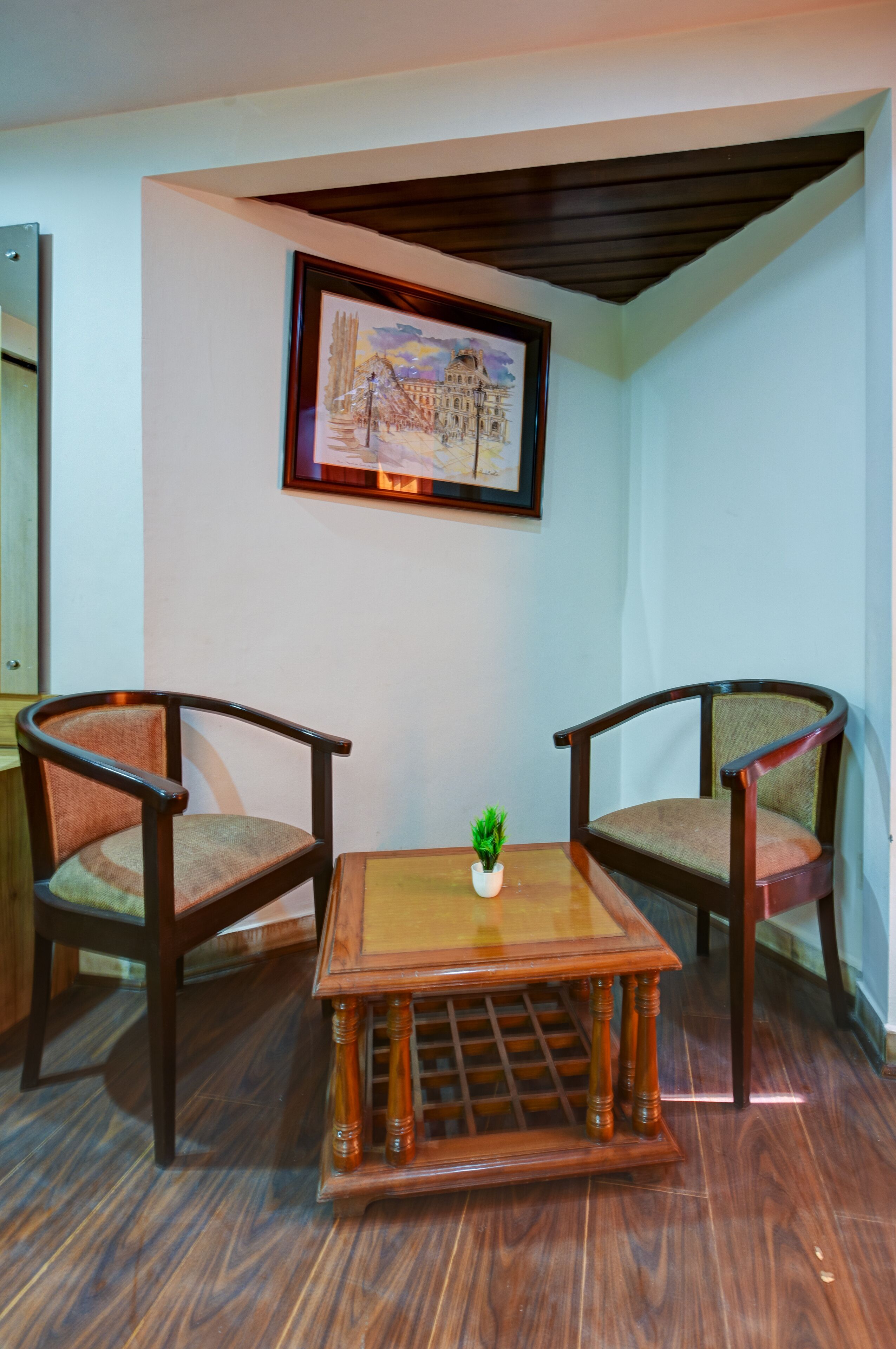 A room with two wooden chairs, a small table with a plant, and a framed picture on the wall.