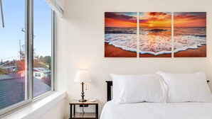 A hotel room with a large window, a bed with white linens, a nightstand with a lamp, and a wall-mounted painting of a beach scene. - Urban Roosevelt/Greenlake Getaway (Seattle)