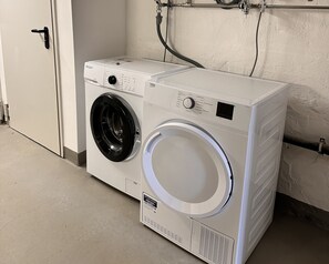 A front-loading washing machine in a laundry room with a door and a wall-mounted hose.